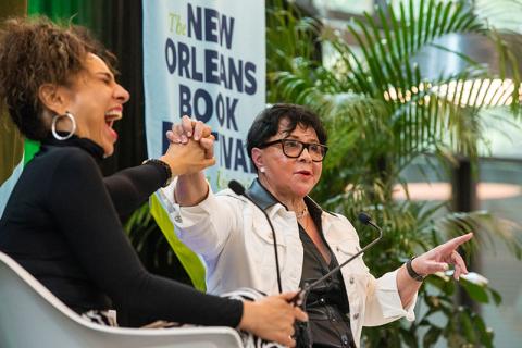 Michelle Miller, wearing large hoop earrings and laughing exuberantly, holds hands with Sheila Johnson during a lively conversation on stage at the New Orleans Book Festival. Sheila Johnson, wearing glasses and a white jacket with black accents, gestures expressively with her other hand as she speaks. A banner in the background reads "The New Orleans Book Festival." Both women are animated, with green plants framing the stage, contributing to the vibrant atmosphere.