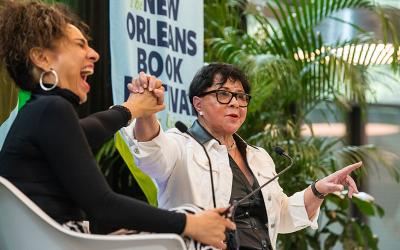 Michelle Miller, wearing large hoop earrings and laughing exuberantly, holds hands with Sheila Johnson during a lively conversation on stage at the New Orleans Book Festival. Sheila Johnson, wearing glasses and a white jacket with black accents, gestures expressively with her other hand as she speaks. A banner in the background reads "The New Orleans Book Festival." Both women are animated, with green plants framing the stage, contributing to the vibrant atmosphere.