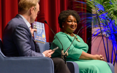 Stacey Abrams in green dress smiles at an interviewer holding her book, "While Justice Sleeps", on stage at the 2024 New Orleans Book Festival at Tulane University.